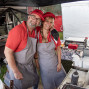 Zwei lächelnde Personen in roten Kappen und grauen Schürzen stehen Arm in Arm an einem Grillstand. Um sie herum sind Grill, Arbeitsflächen, Behälter und Lebensmittel zu sehen, darunter eine belegte Pizza im Vordergrund.