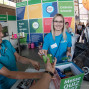 Zwei Mitarbeiterinnen in türkisfarbenen Shirts betreuen einen Messestand der Stadt Schwedt/Oder und geben Besuchern Informationsmaterial. Im Hintergrund sind bunte Werbetafeln, weitere Messebesucher und verschiedene Ausstellungsstände zu sehen.
