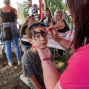 Eine Frau malt einem Jungen bei einer Veranstaltung im Freien ein schwarzes Fledermausmotiv ins Gesicht. Im Hintergrund sitzen und stehen mehrere Erwachsene und Kinder an Tischen unter Bäumen.
