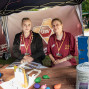 Zwei junge Frauen in bordeauxroten Poloshirts sitzen an einem Informationsstand unter einem Pavillon, hinter ihnen ist ein UBV-Banner zu sehen. Auf dem Tisch liegen Broschüren, Spiel- und Sinnesmaterialien sowie ein Ordner mit der Aufschrift „Kneipp“.