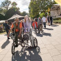 Auf einem sonnigen Platz bei einer Veranstaltung fährt ein Mann ein Lastenfahrrad, in dem ein Mädchen vorne mitfährt. Im Hintergrund sind viele Menschen, Sitzbänke, Zelte und eine kleine Bühne zwischen Bäumen zu sehen.