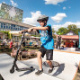 Ein Junge mit Helm fährt auf einem Stunt-Scooter über eine kleine Rampe bei einer Outdoor-Veranstaltung. Im Hintergrund sitzen Menschen an Bierbänken vor einem modernen Gebäude, während weitere Stände und ein blauer Himmel mit Wolken zu sehen sind.
