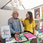 Eine Frau in gelbem Poloshirt interviewt einen älteren Mann mit Mikrofon an einem Informationsstand von viaTEATRI. Auf dem Tisch und an den Wänden sind zahlreiche Broschüren, Plakate und Programmhefte zu Theaterveranstaltungen ausgestellt.
