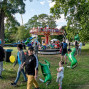 Auf einer grünen Wiese in einem Park steht ein bunt beleuchtetes Kinderkarussell, umgeben von vielen Familien und Kindern. Mehrere Menschen laufen mit Luftballons herum, während große Bäume die fröhliche Jahrmarktsszene einrahmen.