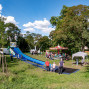 Auf einer grünen Wiese in einem Park stehen mehrere Fahrgeschäfte, darunter eine große blaue Rutsche und ein kleines Karussell. Familien mit Kindern spazieren zwischen den Attraktionen, während hohe Bäume und ein blauer Himmel mit weißen Wolken die Szene umrahmen.