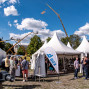 Auf einem Außengelände stehen mehrere weiße Messezelte mit KSB-Fahnen, während Besucher zwischen den Ständen und Ausstellungsstücken umhergehen. Im Hintergrund ragen Arbeitsbühnen und ein großer Kran in den blauen Himmel mit weißen Wolken.