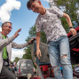 Ein älterer Mann lächelt und hebt die Hand, während ein junger Mann aus einem gekippten orangefarbenen Fahrzeug steigt. Im Hintergrund sind Bäume, ein blauer Himmel mit Wolken und ein Transporter der Verkehrswacht bei einer Veranstaltung zu sehen.