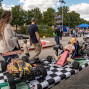 Auf einem öffentlichen Fest stehen Modellrennwagen und ein kleines Go-Kart mit einer Puppe auf einem schwarz-weiß karierten Tisch. Im Hintergrund schauen Familien mit Kindern zu, während Zelte, Fahrzeuge und Bäume die lebhafte Veranstaltung umrahmen.