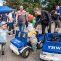 Auf einem belebten Außengelände mit blauen Zelten und THW-Fahrzeugen spielen Kinder mit kleinen blauen Spielzeugautos. Erwachsene stehen daneben, unterhalten sich und helfen den Kindern, während im Hintergrund weitere Besucher und Informationsstände zu sehen sind.