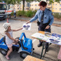 Ein Mädchen sitzt in einem blauen Spielzeugfahrzeug des Technischen Hilfswerks und nimmt von einer Polizistin ein Informationsblatt entgegen. Im Hintergrund steht ein Fahrzeug des Deutschen Roten Kreuzes, während auf einem Tisch Broschüren und Materialien zur Verkehrserziehung ausliegen.