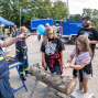 Bei einer Veranstaltung des Technischen Hilfswerks bearbeiten Kinder unter Anleitung einen Baumstamm mit Werkzeugen. Im Hintergrund stehen blaue THW-Fahrzeuge, Zelte und mehrere Besucher auf einem Gelände mit Bäumen.