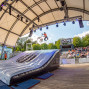Ein BMX-Fahrer springt bei einer Show hoch über ein großes Luftkissen auf einer überdachten Freilichtbühne. Rechts sitzt ein Publikum auf den Tribünen und beobachtet die Vorführung.