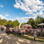 Auf einer großen Außenveranstaltung sitzen viele Menschen an Biertischen unter Sonnenschirmen und Zelten in einem grünen Park. Der Himmel ist blau mit weißen Wolken, und auf dem gepflasterten Weg laufen Besucher, darunter Familien und Personen mit Kinderwagen.
