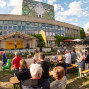 Vor einem modernen Gebäude mit großer Glasfassade findet eine Freiluftveranstaltung mit Bühne, Zelten und vielen Zuschauern statt. Die Menschen sitzen und stehen auf Bierbänken im Sonnenschein, während ein Moderator oder Künstler auf der Bühne spricht.