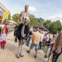 Auf einem belebten Festgelände läuft ein Mann in einem großen Straußenkostüm durch die Menge und zieht die Aufmerksamkeit der Besucher auf sich. Rundherum stehen Familien, Kinder und Stände vor einem Gebäude bei sonnigem Wetter.