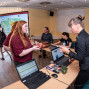 Auf dem Foto sieht man eine Frau in Schwarz, die einer anderen Frau in Rot etwas anbietet, während im Hintergrund mehrere Personen an Tischen sitzen und Laptops benutzen. Es wirkt wie eine Veranstaltung oder ein Messebereich mit Informationsmaterialien und Technik.