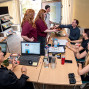 Einige Menschen sitzen an einem großen Tisch, umgeben von Flyern und einem Laptop, während ein Mann einem anderen die Hand schüttelt. Es scheint sich um eine Diskussion oder Präsentation in einer professionellen Umgebung zu handeln.