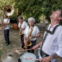 Eine kleine Musikgruppe spielt im Freien zwischen Bäumen, mit Saxophonen, einer Tuba und einem Schlagzeug. Der Schlagzeuger lacht ausgelassen, während die anderen Musiker konzentriert ihre Instrumente spielen.