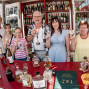 Eine Gruppe von sechs Personen steht an einem Wein- und Spirituosenstand und hält kleine Gläser zum Anstoßen in die Kamera. Vor ihnen liegen zahlreiche Flaschen auf dem Tresen, während im Hintergrund Regale mit Weinflaschen und ein sonniges Festgelände zu sehen sind.