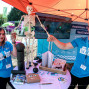 Zwei junge Erwachsene in blauen „Laufen in Aktion“-Shirts stehen an einem Informationsstand unter einem orangefarbenen Pavillon und halten ein Skelettmodell. Auf dem Tisch liegen Gesundheitsbroschüren, Übungsgeräte und Werbematerialien, während im Hintergrund weitere Messestände zu sehen sind.