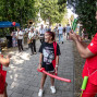 Auf einem belebten Gehweg bei einer Veranstaltung spielt eine kleine Blaskapelle, während Besucher zwischen Ständen und Bäumen umhergehen. Im Vordergrund interagiert ein Mann im roten Anzug mit stacheliger Perücke spielerisch mit zwei Mädchen, von denen eines einen langen roten Ballon hält.
