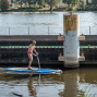Ein Mann in roter Badehose fährt auf einem blauen Stand-up-Paddleboard neben einer Schleusenwand über das Wasser. Im Hintergrund sind der Fluss, Bäume und ein kleines Haus am Ufer zu sehen.