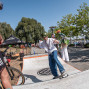 Ein Junge mit Helm fährt auf einem Skateboard über eine kleine Rampe, während ein Mann mit Fahrrad am Rand zuschaut. Im Hintergrund sind Bäume, ein Flussufer, Sitzbänke und eine bunt verkleidete Stelzenfigur zu sehen.