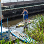 Eine Frau in einem blauen Kleid steht auf einem Stand-up-Paddleboard im Wasser nahe einer Schleuse, während Schilf am Ufer wächst. Links steigt ein Mann in roter Badehose über eine Plattform ins Wasser, im Hintergrund liegt ein großes dunkles Schiff.