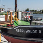 Das Foto zeigt ein festgemachtes Schiff mit dem Namen „EBERSWALDE“ an einem Anleger auf einem Fluss oder Kanal. An Deck sind Rettungsringe, Geländer und eine deutsche Flagge zu sehen, im Hintergrund liegen Wasser und Bäume unter klarem Himmel.