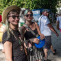 Zwei junge Frauen mit Sonnenbrillen stehen lächelnd auf einem belebten Gehweg, eine trägt einen Strohhut und die andere hält eine blaue Frisbee-Scheibe. Im Hintergrund laufen mehrere Menschen vorbei, daneben sind rote Luftballons und grüne Bäume zu sehen.