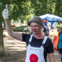 Eine lächelnde Frau mit Hut, Mikrofon und weißer Latzhose hält in einem Park eine kleine Glocke hoch. Im Hintergrund sind Bäume, ein blaues Zelt und mehrere Menschen bei einer Veranstaltung zu sehen.