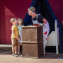Eine verkleidete Schauspielerin mit Hut und Mikrofon beugt sich auf einer Bühne zu zwei Kindern hinunter. Vor ihnen steht eine große alte Truhe, im Hintergrund sind dunkle Vorhänge und ein weißes Bett zu sehen.