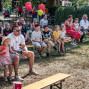 Eine Gruppe von Erwachsenen und Kindern sitzt im Schatten auf Bierbänken bei einer Veranstaltung im Freien. Einige Kinder halten Luftballons, während die Zuschauer aufmerksam nach vorne schauen.