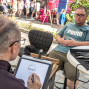 Ein Straßenkünstler zeichnet auf einem Tablet eine Karikatur eines Mannes, der ihm gegenüber auf einem Stuhl sitzt. Auf dem Tisch liegen mehrere Stifte, während im Hintergrund Menschen an Ständen vorbeigehen und Pflanzen die Szene umrahmen.